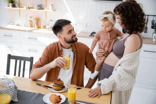 Donna riccia con neonata in braccio guardando il marito sorridente durante la colazione — Foto stock