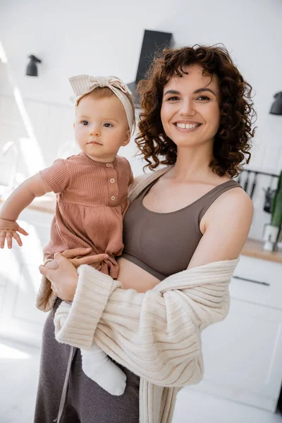 Позитивная мама с кудрявыми волосами улыбается, держа в руках младенца в повязке на голове — стоковое фото