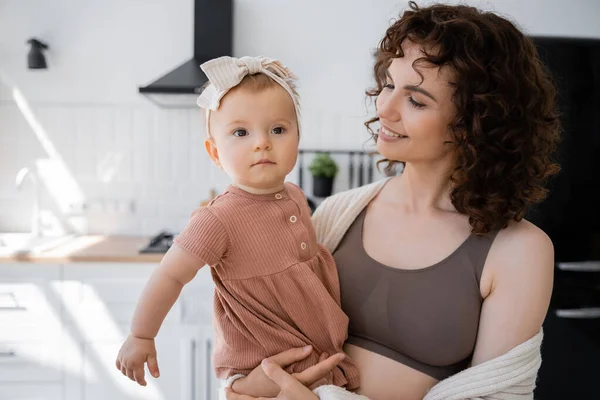 Fröhliche Mutter mit lockigem Haar, die lächelt, während sie das Kleinkind im Stirnband im Arm hält — Stockfoto