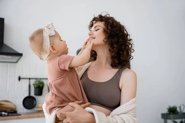 Щаслива жінка з кучерявим волоссям посміхається, тримаючись в руках немовляти в пов'язці — стокове фото