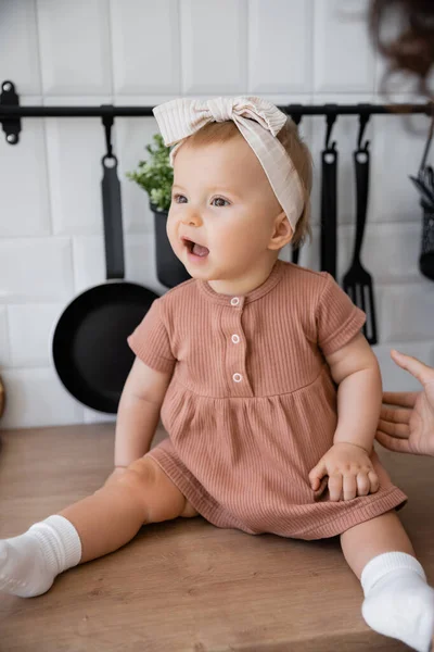 Amazed infant girl in headband sitting on kitchen worktop near blurred mother — Stock Photo