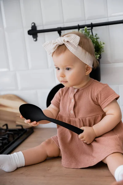 Neonata in fascia e abito rosa che tiene mestolo mentre seduto sul piano di lavoro della cucina — Foto stock