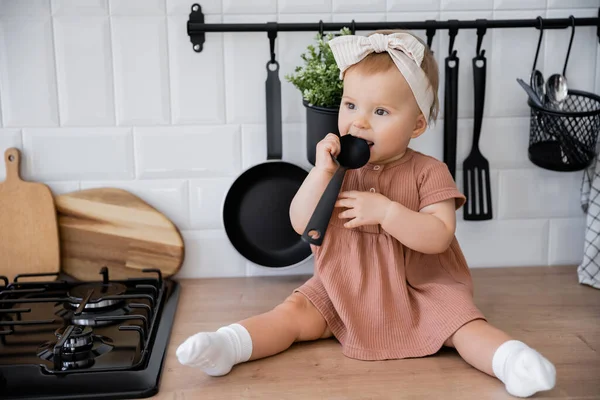 Bébé fille en bandeau et robe rose tenant louche près de la bouche tout en étant assis sur le plan de travail de la cuisine — Photo de stock