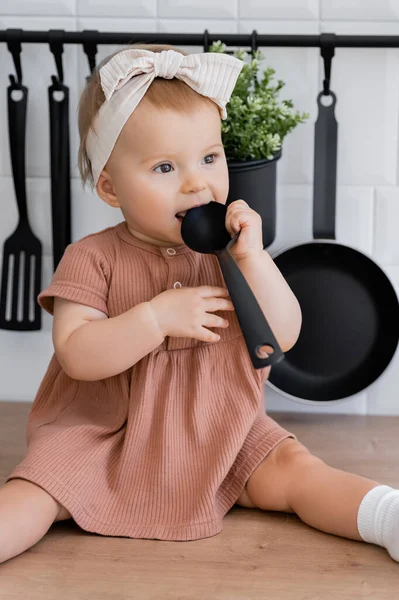 Baby girl in headband and pink dress holding ladle near mouth while sitting on kitchen worktop — Stock Photo