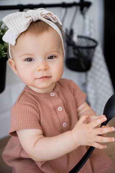 Portrait of infant girl in headband and pink dress holding ladle while sitting on kitchen worktop — Stock Photo