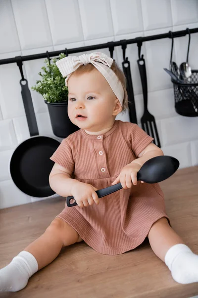 Bébé fille en bandeau et robe rose tenant louche tout en étant assis sur le plan de travail de la cuisine et regardant loin — Photo de stock