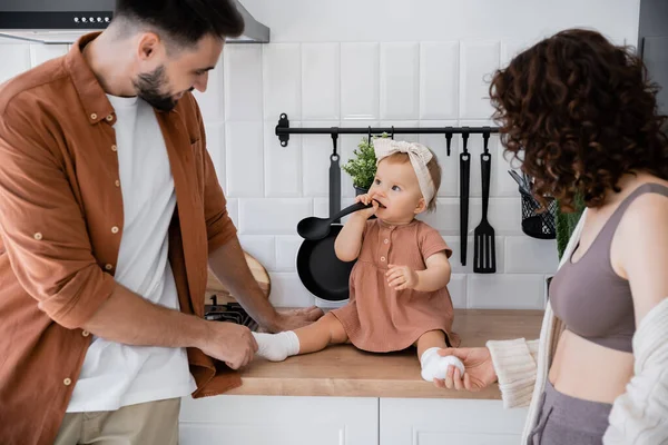 Infant child in headband holding ladle near mouth while sitting on kitchen worktop near parents — Stock Photo