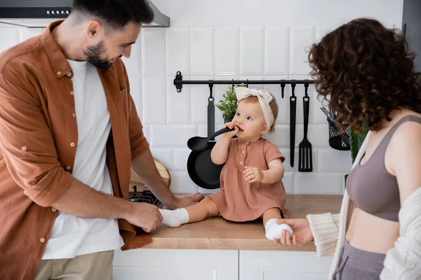 Infant child in headband holding ladle near mouth while sitting on kitchen worktop near parents — Stock Photo