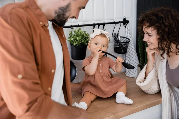 Младенец в повязке держит ковш у рта, сидя на кухонном столе рядом с веселыми родителями — стоковое фото