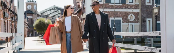 Gioiosa coppia multiculturale in occhiali da sole che trasporta borse della spesa e si tiene per mano sul ponte della città, banner — Foto stock
