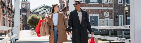Joyful multicultural couple in sunglasses carrying shopping bags and holding hands on city bridge, banner — Stock Photo