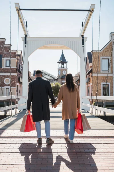 Back view of multiethnic couple in coats walking with shopping bags across bridge and holding hands — Stock Photo