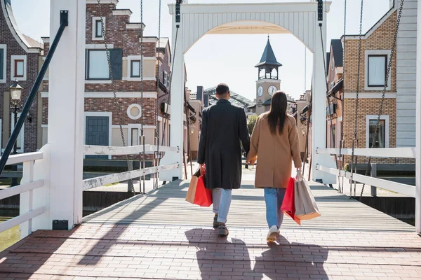 Back view of interracial couple in coats holding hands and walking on bridge with shopping bags — Stock Photo