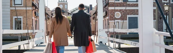 Rückansicht des trendigen multiethnischen Paares mit Einkaufstüten, die Händchen halten auf Brücke, Banner — Stockfoto
