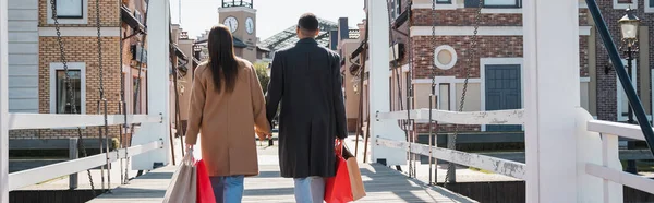 Back view of trendy multiethnic couple with shopping bags holding hands on bridge, banner — Stock Photo