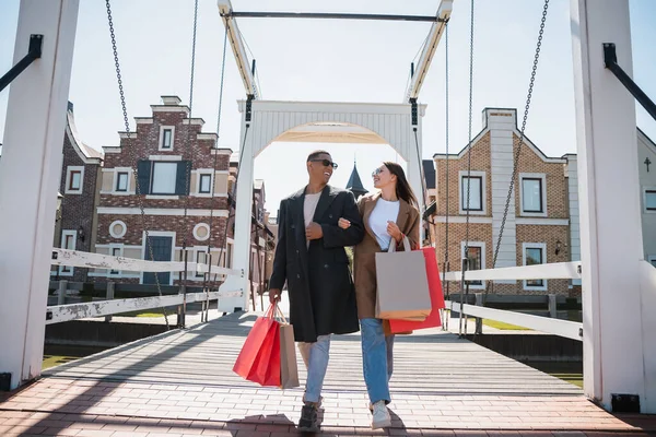 Stilvolles gemischtrassiges Paar lächelt sich an und läuft mit Einkaufstüten über Brücke — Stockfoto