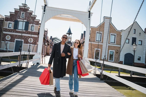 Alegre pareja multiétnica en ropa elegante y gafas de sol caminando con bolsas de compras a través del puente - foto de stock