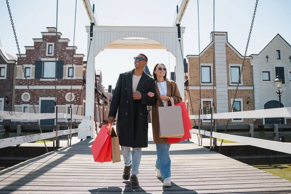 Volle Länge des lächelnden gemischtrassigen Paares in Sonnenbrille und Mantel, das mit Einkaufstaschen auf Brücke läuft — Stockfoto