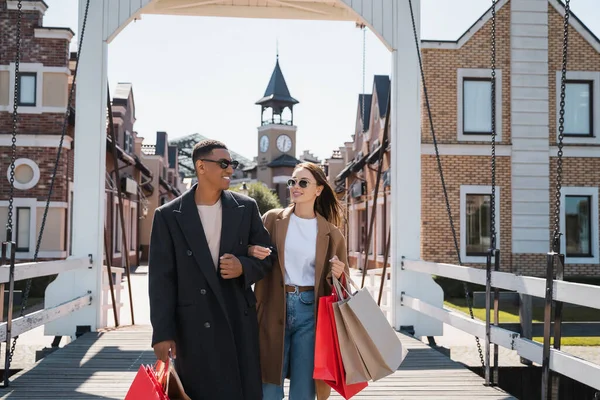 Pareja multiétnica de moda y feliz con bolsas de compras en el puente de la ciudad - foto de stock