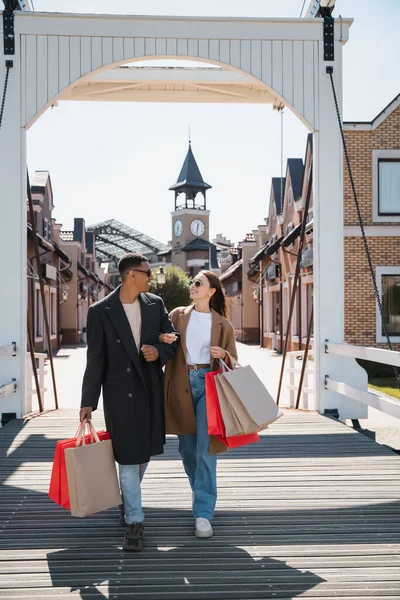 Pareja multiétnica de moda en gafas de sol caminando con bolsas de compras y sonriéndose mutuamente en el puente de la ciudad - foto de stock