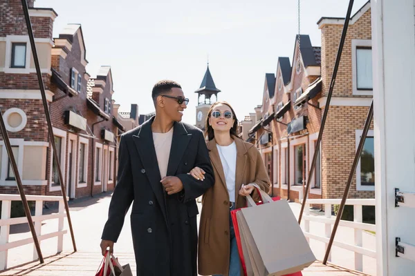 Sonriente afroamericano hombre en gafas de sol mirando a una novia feliz con bolsas de compras en puente urbano - foto de stock