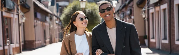 Casal interracial na moda em casacos e óculos de sol sorrindo na rua urbana, bandeira — Fotografia de Stock