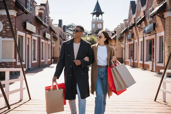 Interracial couple in trendy coats and sunglasses walking with shopping bags and looking away on bridge — Stock Photo