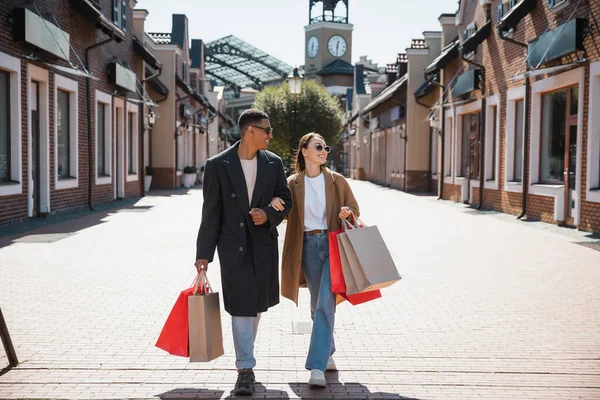 Volle Länge des multiethnischen Paares in Mänteln und Sonnenbrillen, das mit Einkaufstüten auf der städtischen Straße spaziert — Stockfoto