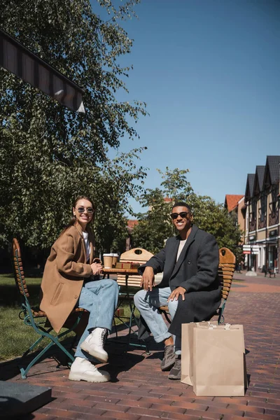 Glückliches multiethnisches Paar in Mänteln und Sonnenbrillen, das neben Coffee to go sitzt und im Straßencafé Taschen einkauft — Stockfoto