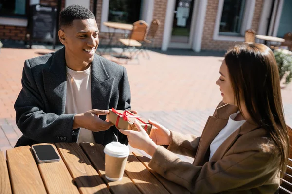 Élégant homme afro-américain présentant cadeau de Noël à la jeune femme pendant la date dans un café de rue — Photo de stock