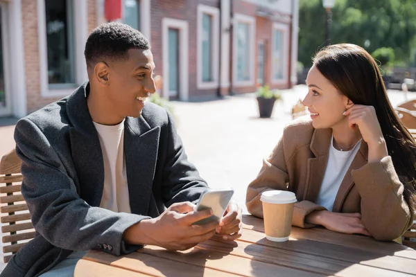 Stilvoller afrikanisch-amerikanischer Mann mit Handy im Gespräch mit einer jungen und lächelnden Frau in der Nähe von Pappbechern in einem Straßencafé — Stockfoto