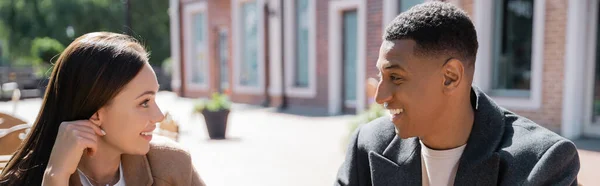 Heureux couple interracial en vêtements à la mode souriant et parlant sur la rue de la ville, bannière — Photo de stock