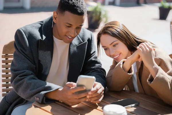 Fröhliches multiethnisches Paar, das auf der Caféterrasse auf sein Handy schaut — Stockfoto