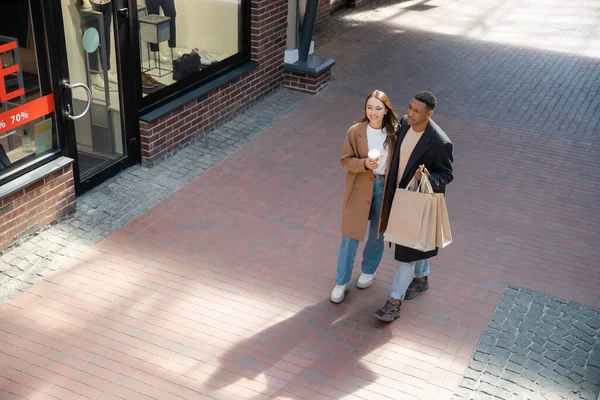Vista de alto ângulo de casal multicultural elegante com sacos de compras e copo de papel andando ao longo da loja com vitrines — Fotografia de Stock