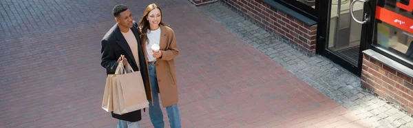 Cheerful interracial couple in stylish coats holding takeaway drink and shopping bags on city street, banner — Stock Photo