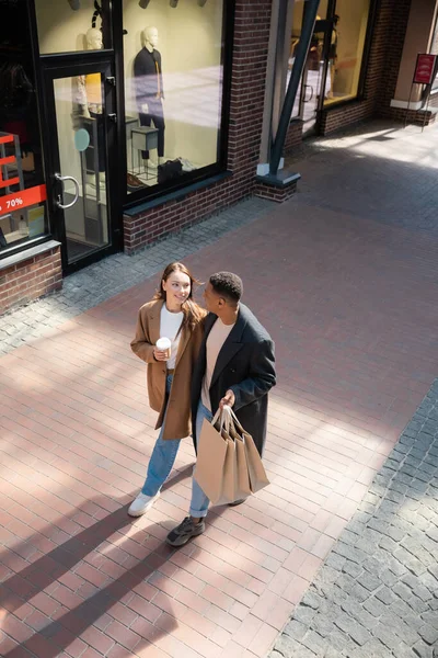 Vista de ángulo alto de la pareja interracial en abrigos de moda caminando con bolsas de compras y café para ir a lo largo de vitrinas - foto de stock