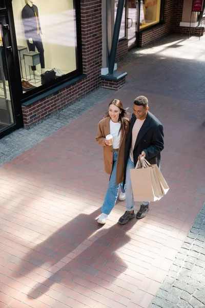 Hochwinkel-Ansicht von glücklichen und modischen multiethnischen Paar mit Einkaufstaschen und Pappbecher zu Fuß in der Nähe von Geschäften auf der Straße — Stockfoto