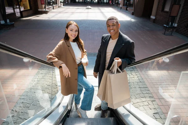 Trendy woman with takeaway drink and african american man with shopping bags smiling at camera on escalator — Stock Photo