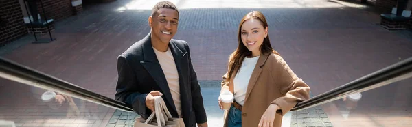 Pareja multiétnica de moda con bolsas de compras y café para ir sonriendo a la cámara en la escalera mecánica, pancarta - foto de stock