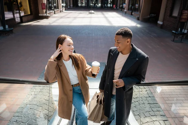 Mulher feliz segurando café para ir perto de homem americano africano na moda com sacos de compras na escada rolante — Fotografia de Stock