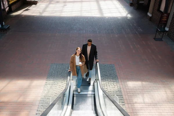 Vista de ángulo alto de la mujer de moda con café para ir y hombre afroamericano con bolsas de compras en escaleras mecánicas - foto de stock