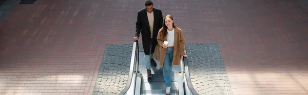 Vista de ángulo alto de la pareja multiétnica en abrigos de moda sosteniendo taza de papel y bolsas de compras en escaleras mecánicas, pancarta - foto de stock