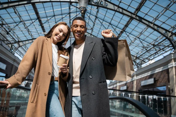 Joyful interracial couple in trendy coats holding shopping bags and coffee to go while smiling at camera under glass roof — Stock Photo