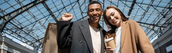 Trendiges multiethnisches Paar mit Einkaufstaschen und Imbissgetränk, lächelnd in die Kamera, Banner — Stockfoto