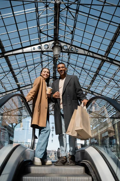 Mujer de moda con taza de papel y hombre afroamericano con bolsas de compras sonriendo a la cámara en escaleras mecánicas bajo techo de cristal - foto de stock