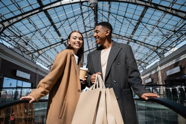 Mujer alegre con café para ir mirando a la cámara cerca de hombre afroamericano con bolsas de compras - foto de stock