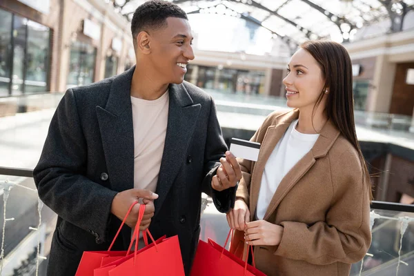 Glücklicher afrikanisch-amerikanischer Mann zeigt seiner zufriedenen jungen Freundin mit Einkaufstüten seine Kreditkarte — Stockfoto