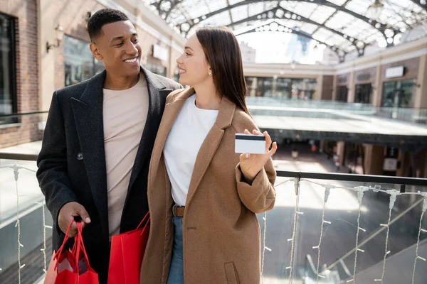 Donna alla moda in possesso di carta di credito vicino sorridente fidanzato africano americano con borse della spesa — Foto stock