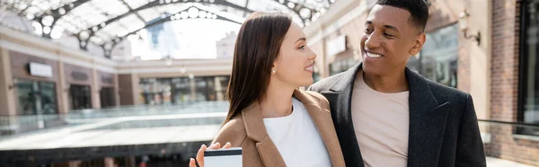 Feliz joven con tarjeta de crédito cerca de novio afroamericano de moda, pancarta - foto de stock