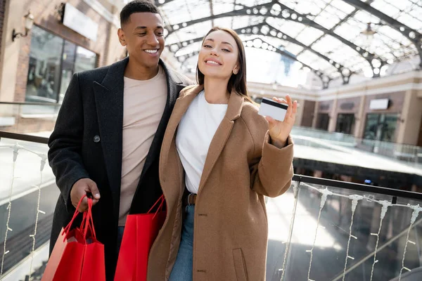 Jeune femme avec carte de crédit souriant à la caméra près élégant homme afro-américain avec des sacs à provisions — Photo de stock